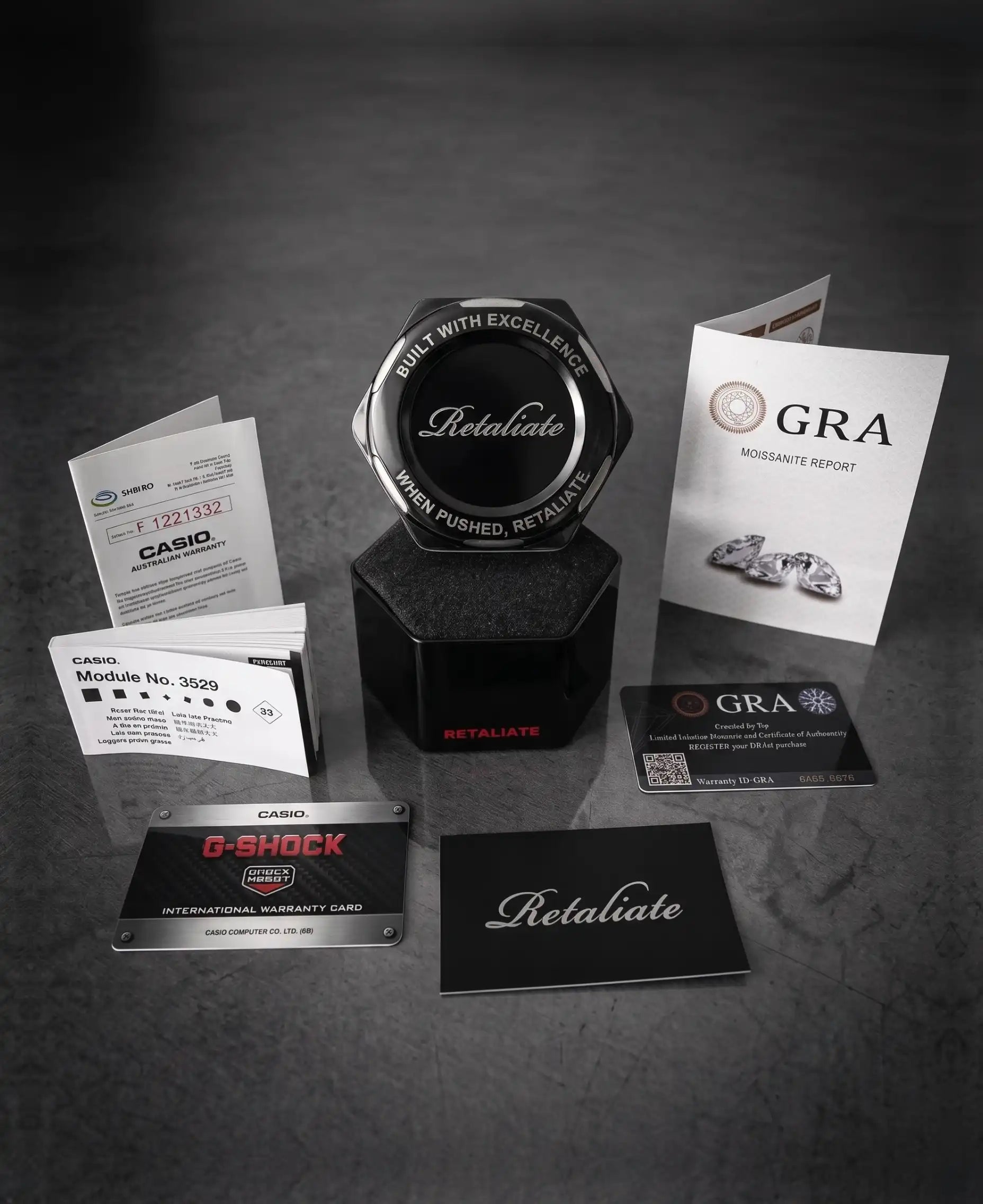 A jewelry service kit featuring a central black hexagonal display stand with the word 'Retaliate' on it, surrounded by branded business cards, a CASIO G-Shock watch, a diamond report card, and informational brochures on a dark textured background.